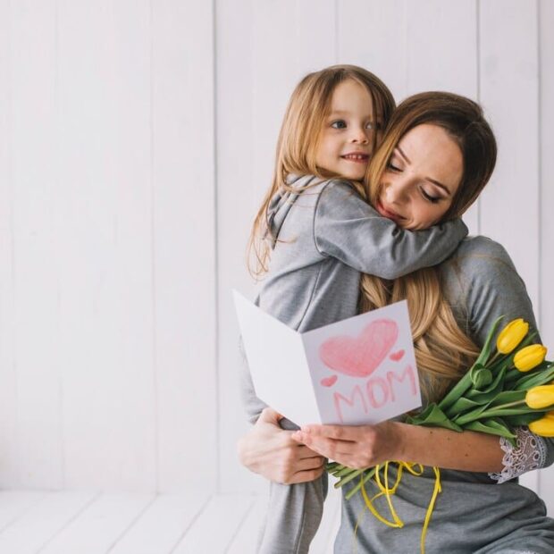 Mãe segura filha pequena no colo junto a um ramalhete de rosas amarelas, enquanto lê um cartão.