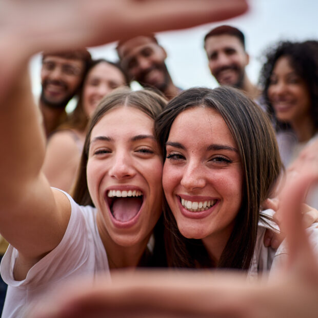 Duas amigas sorrindo fazem foto, com enquadramento dos polegares, e amigos ao fundo conversando