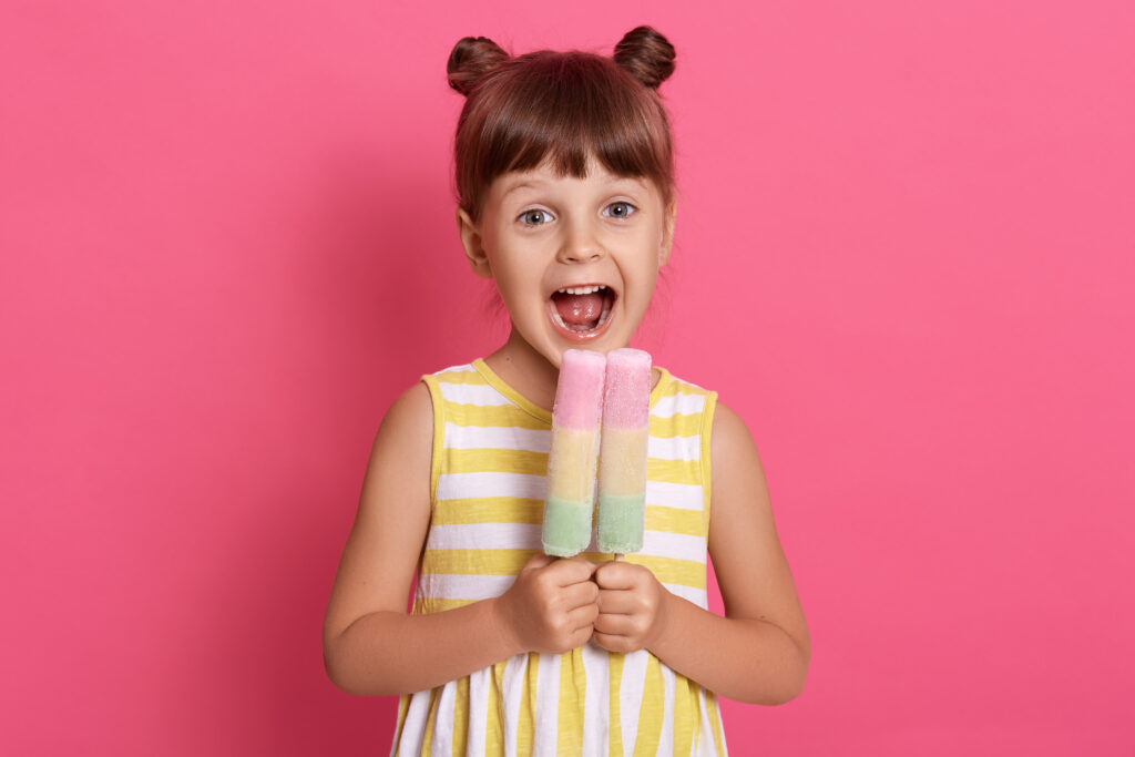 small european kid biting fruit ice cream, little charming girl with widely opened mouth, wearing summer clothes, looks happy, having fun with delicious dessert.