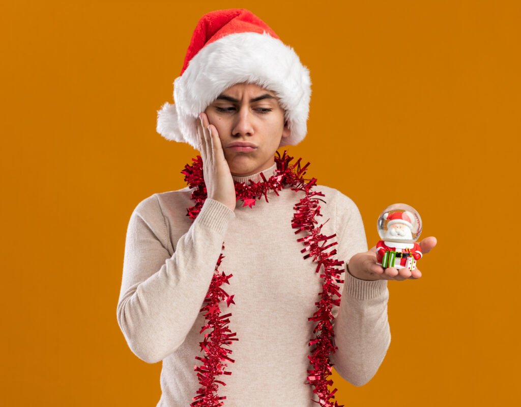 thinking young guy wearing christmas hat with garland on neck holding and looking at christmas toy putting hand on cheek isolated on yellow background