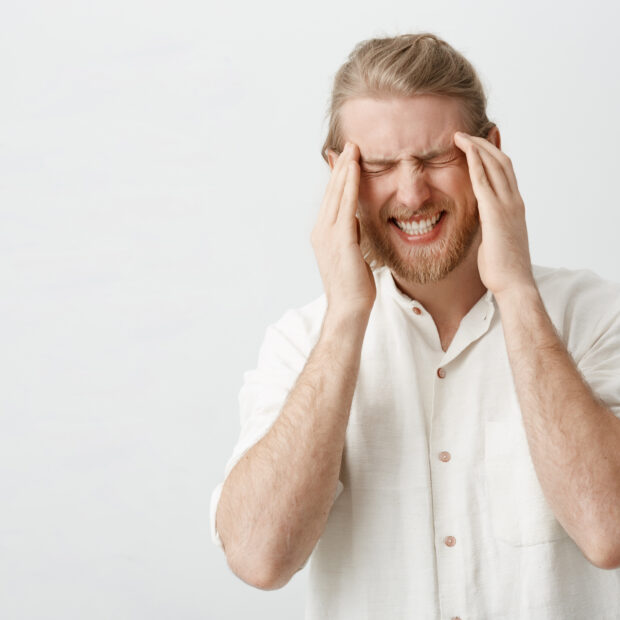indoor portrait of caucasian blond man with beard suffering from migraine or headache, holding fingers on temples, frowning and squeezing teeth from pain, standing over gray background.