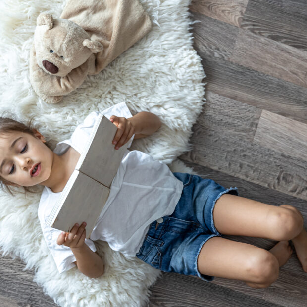 little girl reads a book lying on the floor at home, top view.