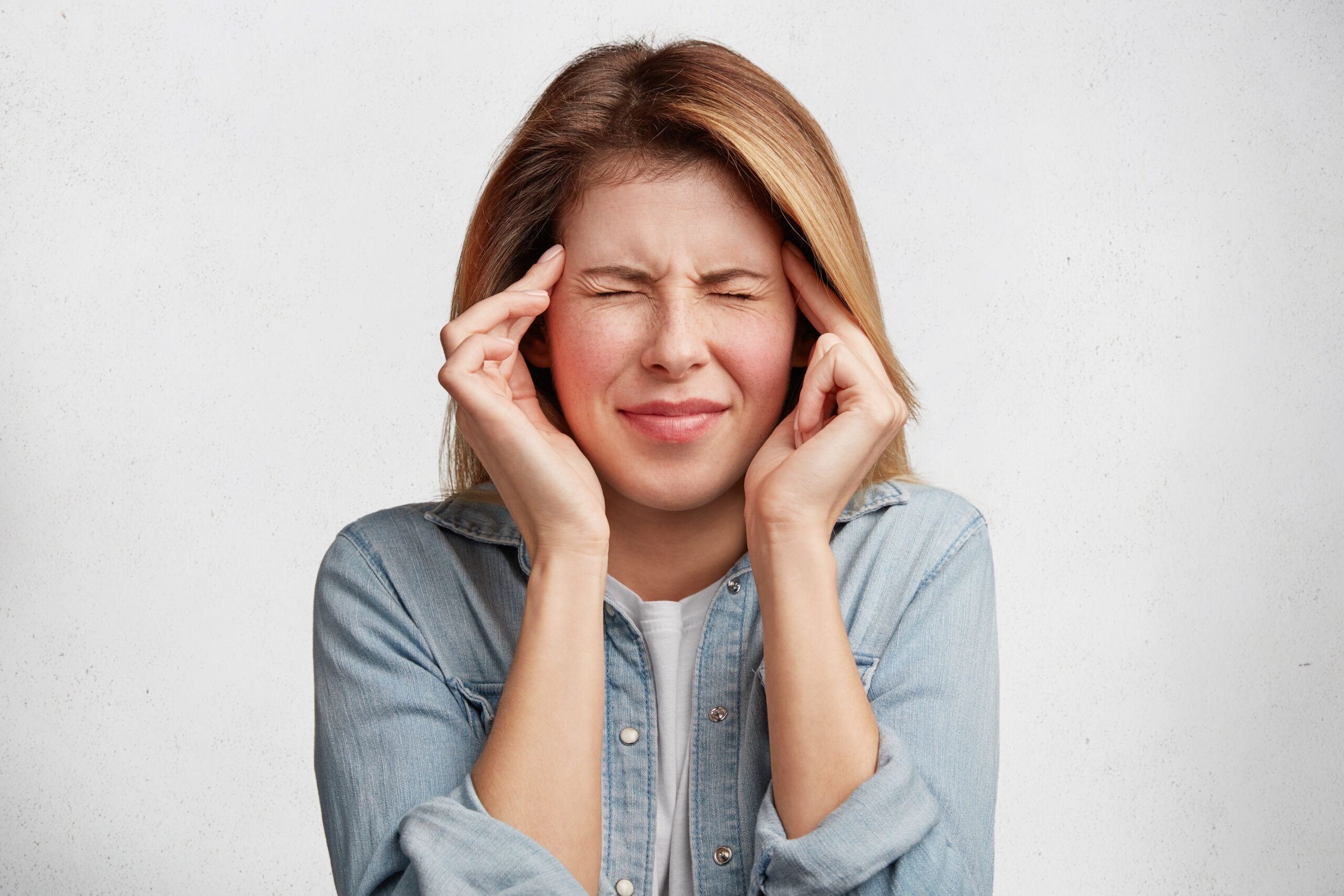 indoor shot of depressed female model wears denim jacket, keeps finger on temples, suffers from headache, feels pain, being overworked, need rest and calm. people, tiredness, exhaustion concept