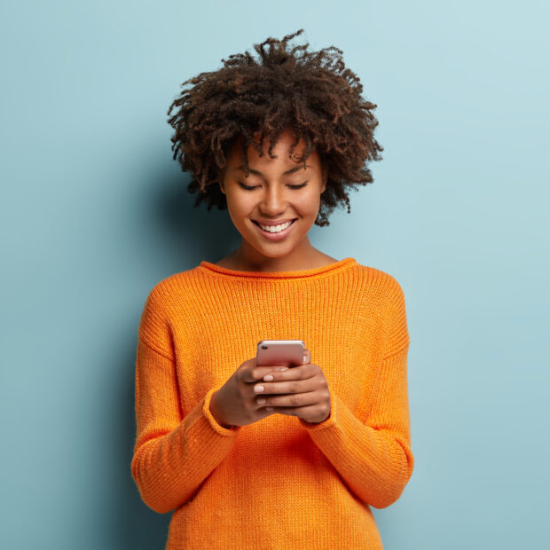 satisfied hipster girl with afro haircut, types text message on cell phone, enjoys online communication, types feedback, wears orange jumper, isolated on blue studio wall. technology concept