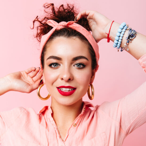 close up portrait of blue eyed lady with red lipstick dressed in pink shirt and massive earrings on isolated background.