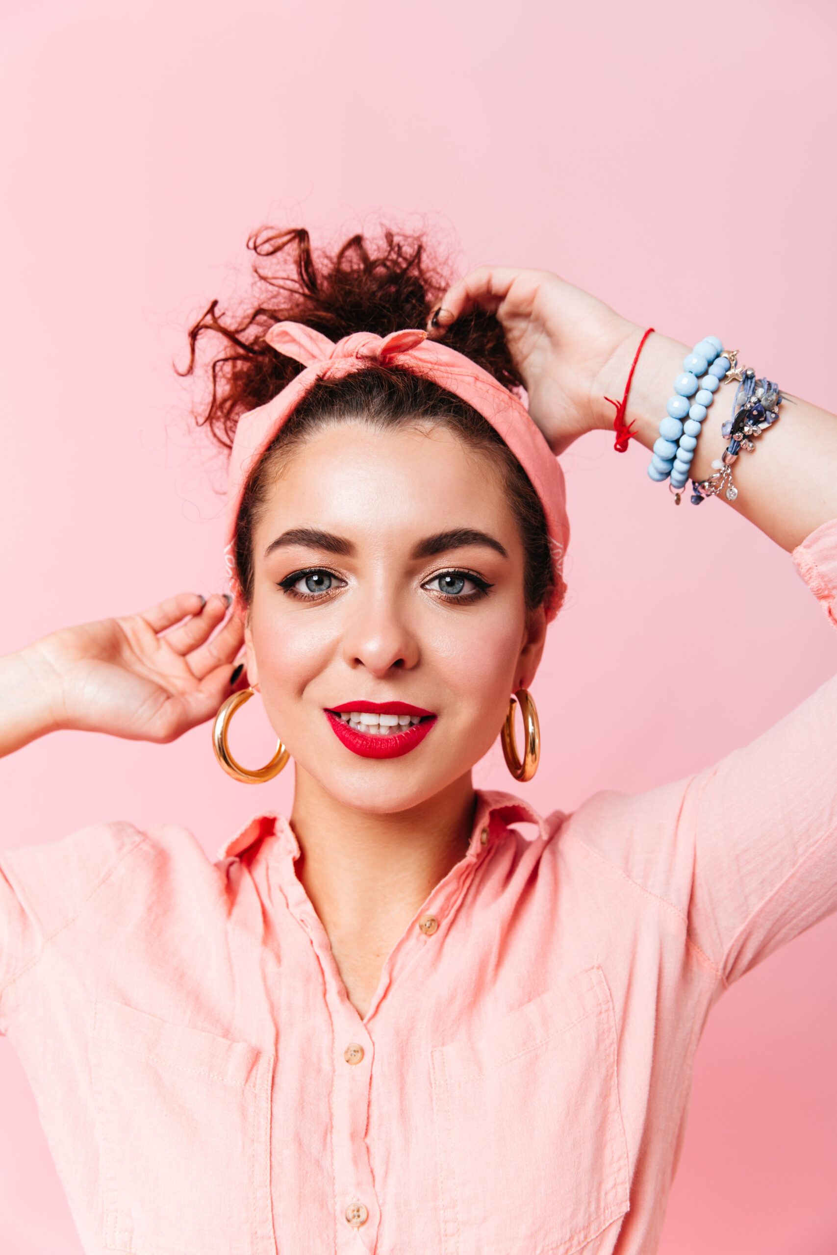close up portrait of blue eyed lady with red lipstick dressed in pink shirt and massive earrings on isolated background.