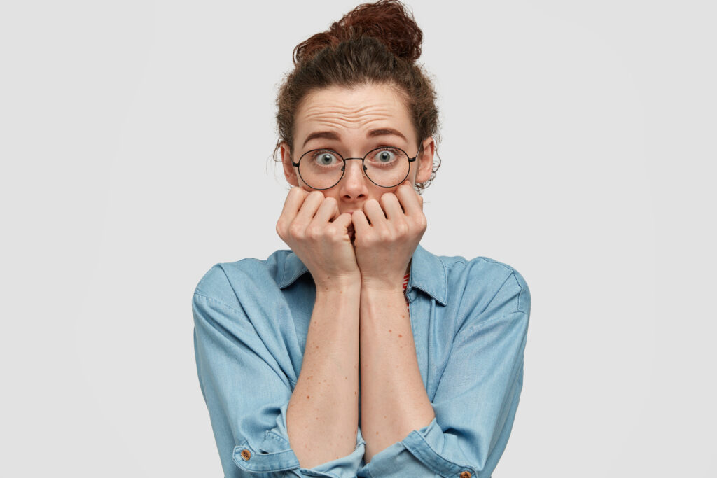 lovely blue eyed female feels scared of ghosts at night, bites fingernails with fear, feels insecure, wears spectacles and denim jacket, isolated over white background. people and anxiety concept