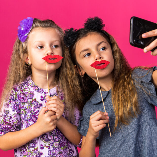 pleased little pretty girls holding fake lips on sticks taking selfie isolated on pink background with copy space
