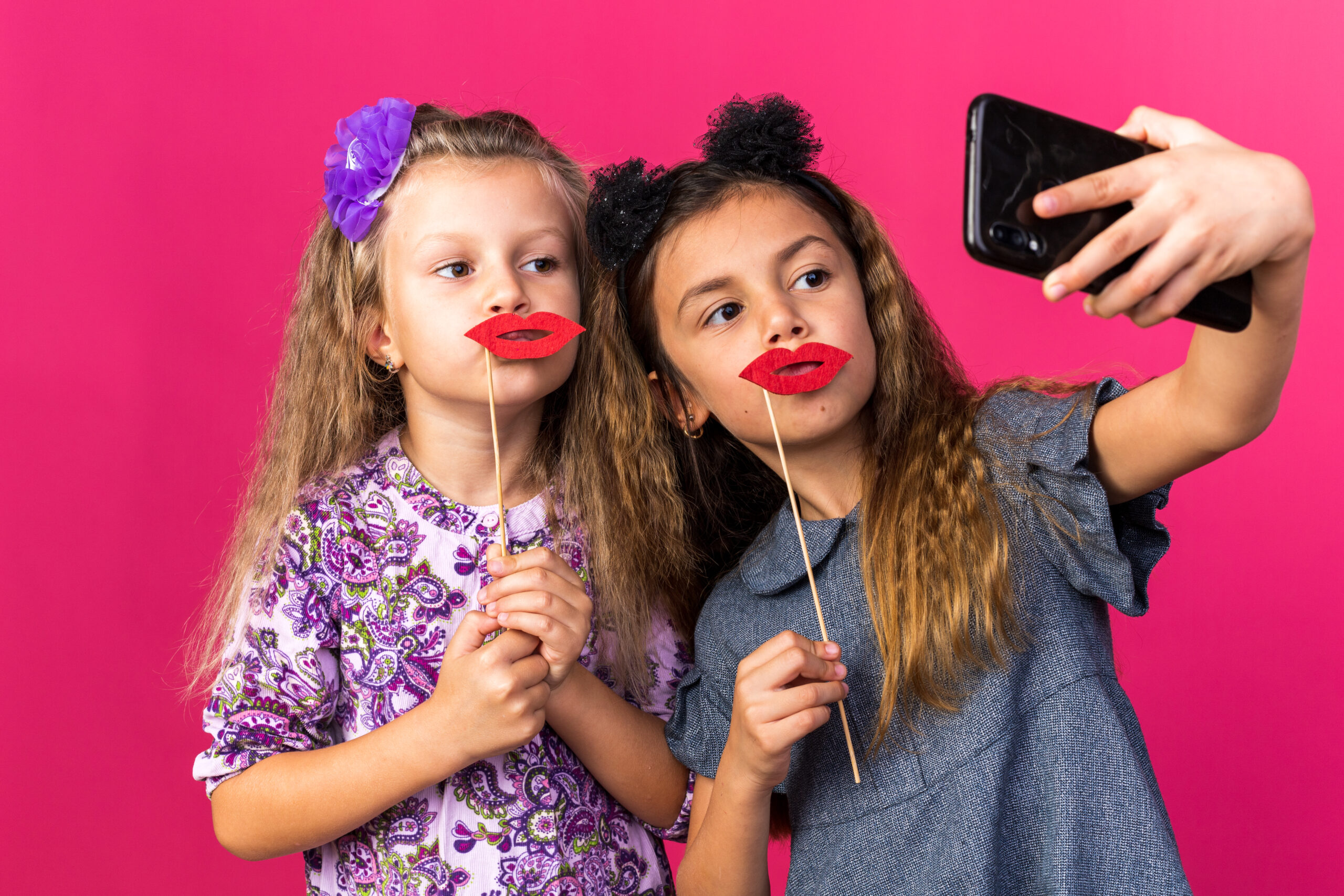 pleased little pretty girls holding fake lips on sticks taking selfie isolated on pink background with copy space