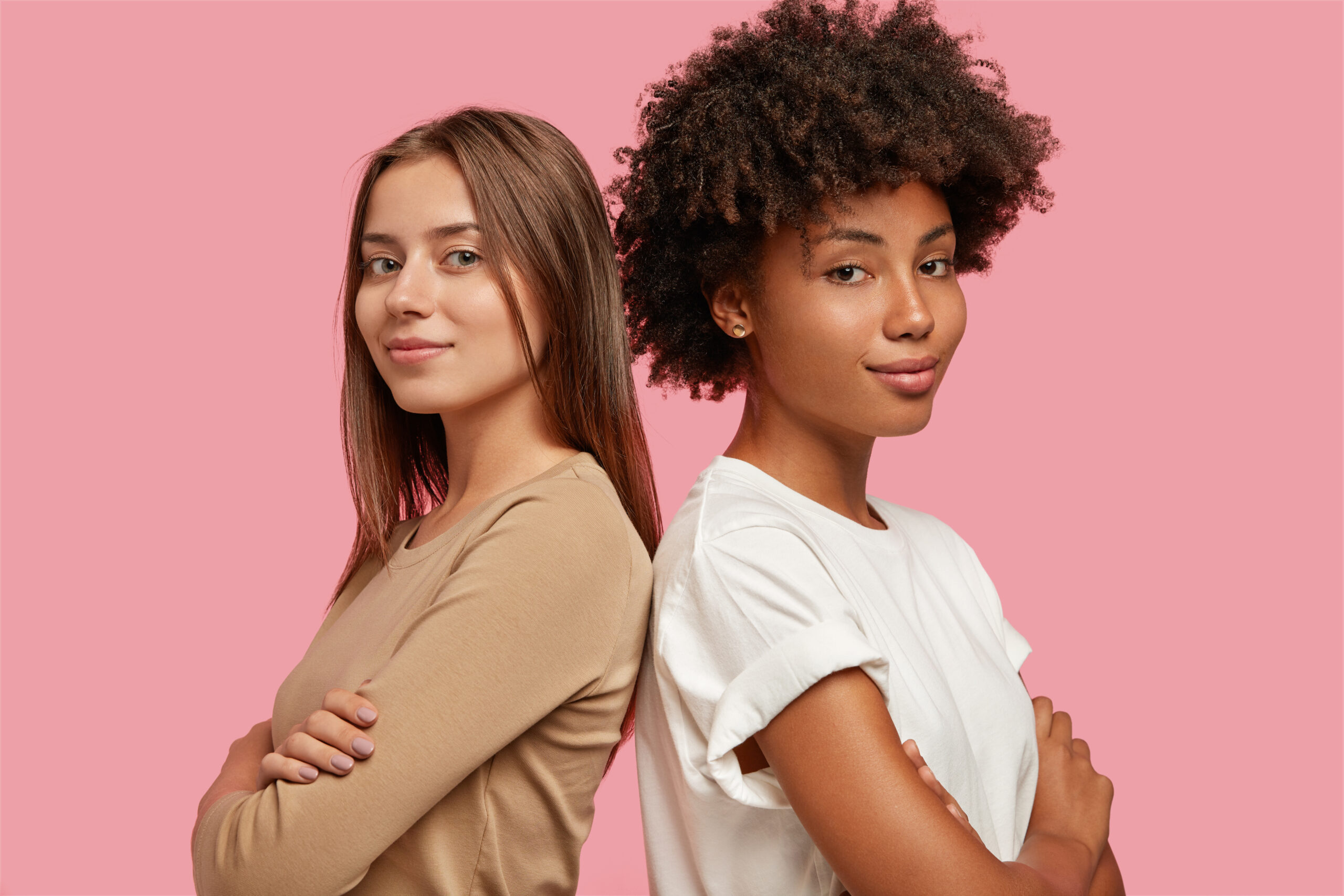 diversity and feminity concept. confident young mixed race women stand backs to each other, keep hands crossed over chest, collaborate for common task, dressed casually, isolated over pink background.