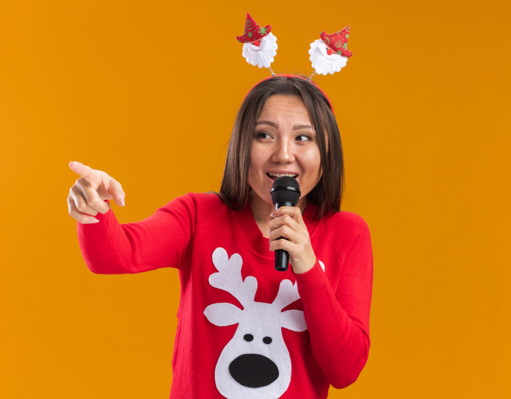 smiling young asian girl wearing christmas hair hoop with sweater speaks on microphone points at side isolated on orange background