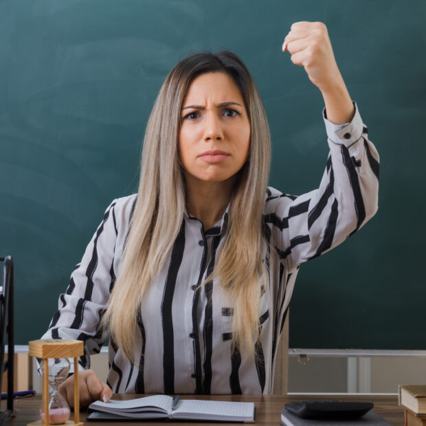 young woman teacher sitting at school desk in front of blackboard in classroom checking homework of students angry and mad raising clenched fist