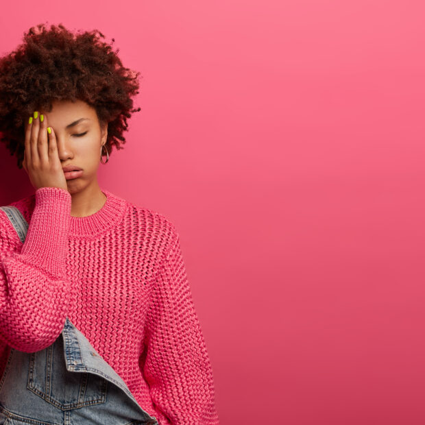 dissatisfied tired woman covers face from exhaustion, closes eyes, sighs deeply, wears loose jumper and overalls, has headache after long working hours, isolated on pink background, copy space area