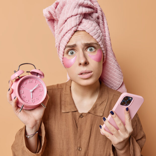 worried young woman awakes late holds alarm clock and smartphone undergoes morning routines dressed in pajama wrapped towel on head isolated over brown background. awakening and rest concept