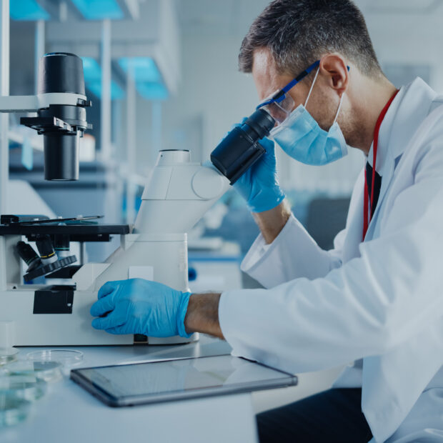 medical development laboratory: scientist wearing face mask looking under microscope and using digital tablet. specialists working on medicine, biotechnology research in advanced pharma lab