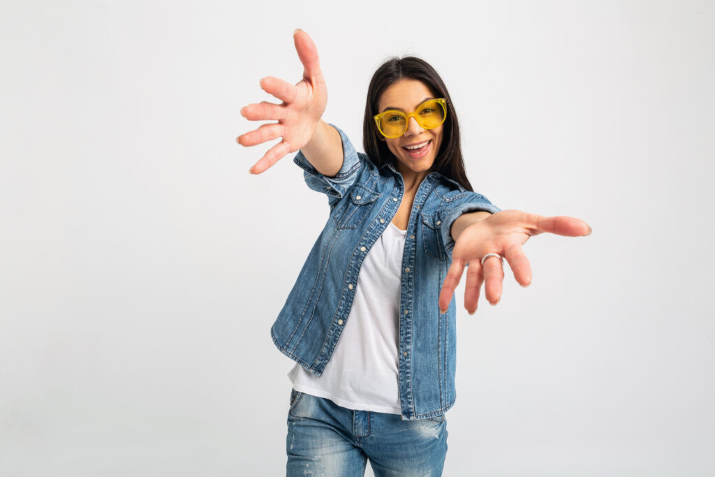 attractive laughing emotional woman open arms to camera isolated on white studio background dressed jeans and denim shirt wearing yellow sunglasses hipster style outfit having fun