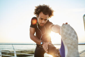 sporty afro american boy in black sportwear and blue sneakers stretching his muscles straightening his leg against plarform, preparing for jogging workout. young dark skinned man jogger warming up before run.