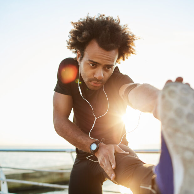 sporty afro american boy in black sportwear and blue sneakers stretching his muscles straightening his leg against plarform, preparing for jogging workout. young dark skinned man jogger warming up before run.