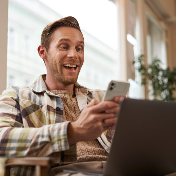 image of man in cafe, coffee shop visitor sitting in chair with laptop and smartphone, looking surprised and excited at mobile phone screen, amazed by big news, promo offer on app
