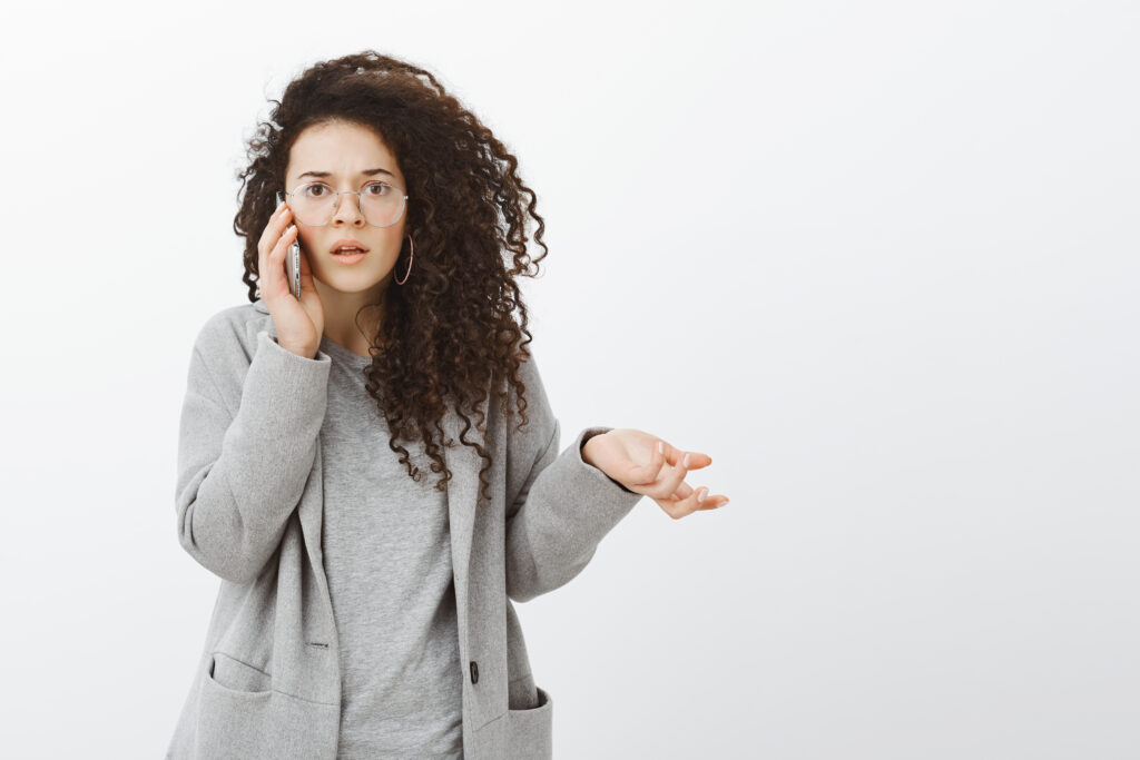 studio shot of confused clueless caucasian businesswoman in trendy eyewear and coat, raising palm questioned and staring frustrated at camera, standing shocked over gray background