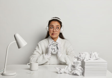 horizontal shot of thoughtful unaware student bites lips and looks hesitantly, holds alarm clock, listens music in headset, thinks about exam preparation, works with papers, poses at white desktop