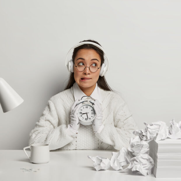 horizontal shot of thoughtful unaware student bites lips and looks hesitantly, holds alarm clock, listens music in headset, thinks about exam preparation, works with papers, poses at white desktop