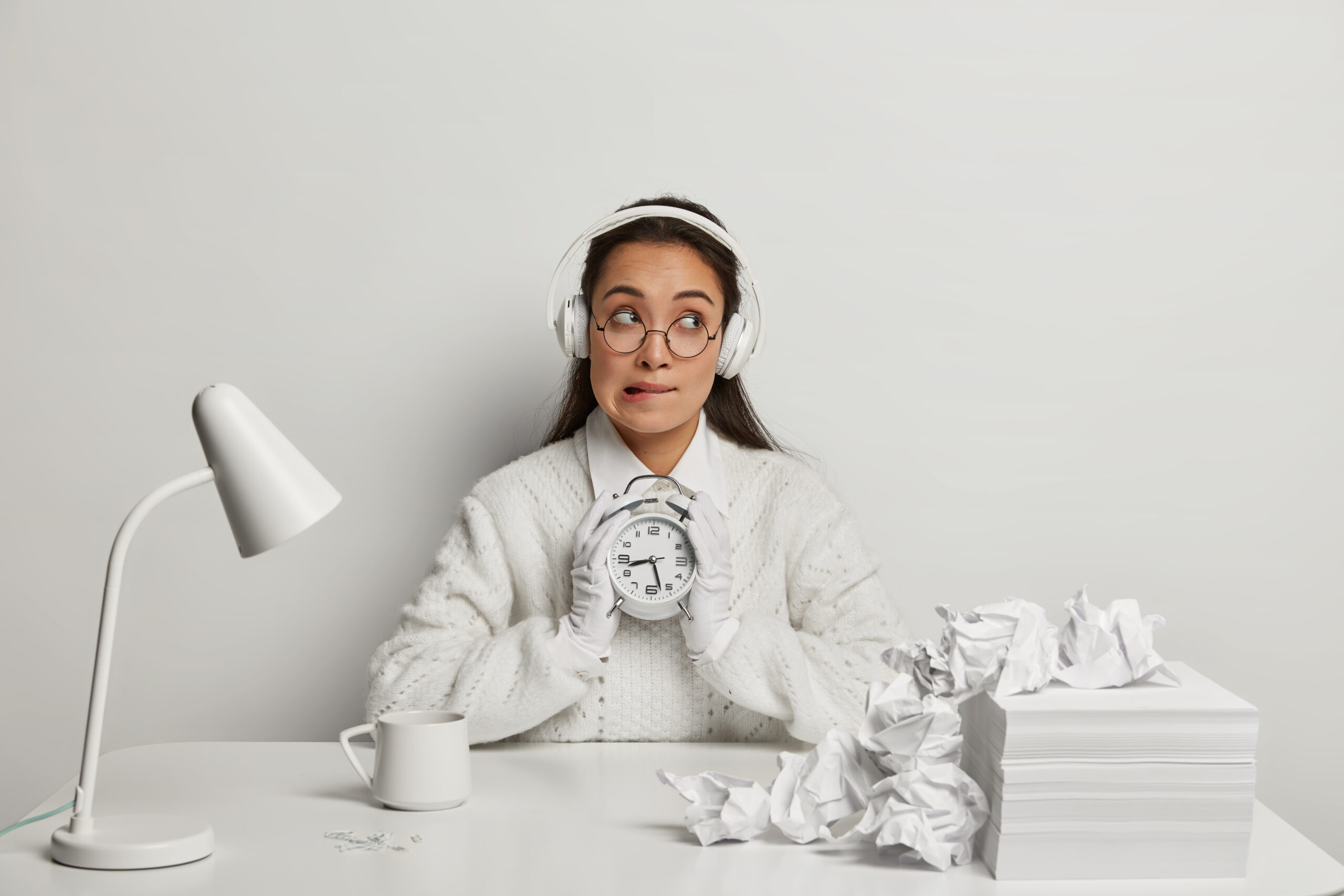 horizontal shot of thoughtful unaware student bites lips and looks hesitantly, holds alarm clock, listens music in headset, thinks about exam preparation, works with papers, poses at white desktop
