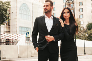 beautiful fashion woman and her handsome elegant boyfriend in suit. sexy brunette model in black evening dress. fashionable couple posing in the street in europe. brutal man and his female outdoors