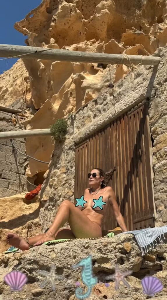 Atriz aparece nua em uma cena na praia, sentada na areia e cobrindo os seios