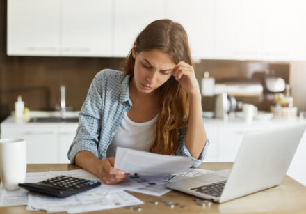 attractive young housewife wearing shirt at home studying gas and electricity bills, checking calculations, looking at sheet of paper in her hands with serious and focused expression on her face