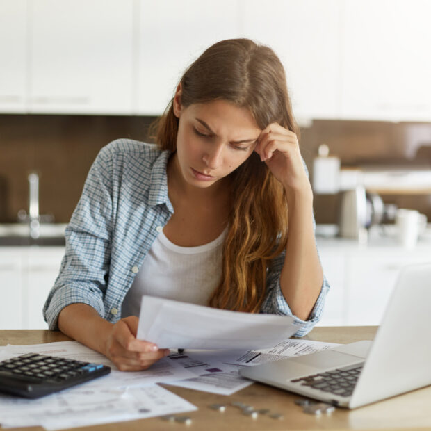 attractive young housewife wearing shirt at home studying gas and electricity bills, checking calculations, looking at sheet of paper in her hands with serious and focused expression on her face