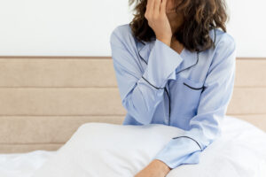 young beautiful woman in blue pajamas sitting on bed with pillow waking up yawning in bedroom interior on light background