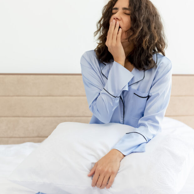 young beautiful woman in blue pajamas sitting on bed with pillow waking up yawning in bedroom interior on light background