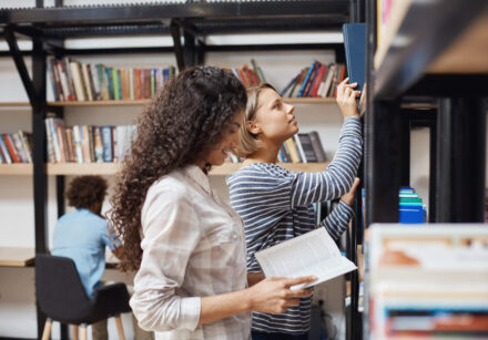 two young cheerful female students in casual clothes standing near bookshelves in university library looking through literature for team project.