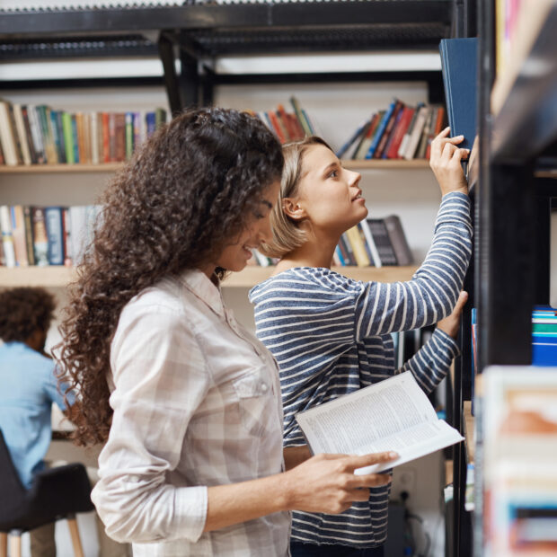 two young cheerful female students in casual clothes standing near bookshelves in university library looking through literature for team project.