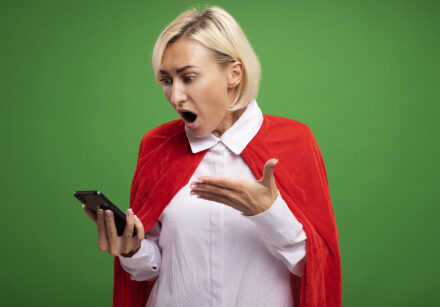 surprised middle aged blonde superhero woman in red cape holding and looking at mobile phone pointing at it with hand isolated on green background with copy space