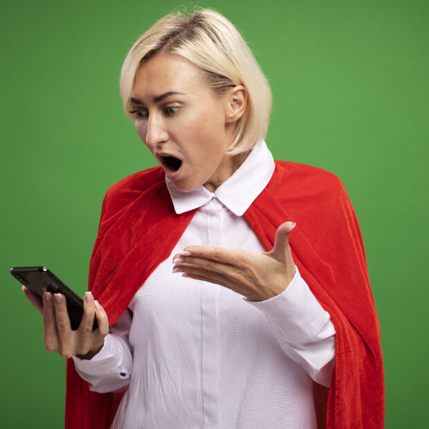 surprised middle aged blonde superhero woman in red cape holding and looking at mobile phone pointing at it with hand isolated on green background with copy space