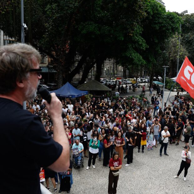 Um homem de costas fala ao microfone para uma multidão de manifestantes em uma praça urbana com uma bandeira vermelha ao fundo.