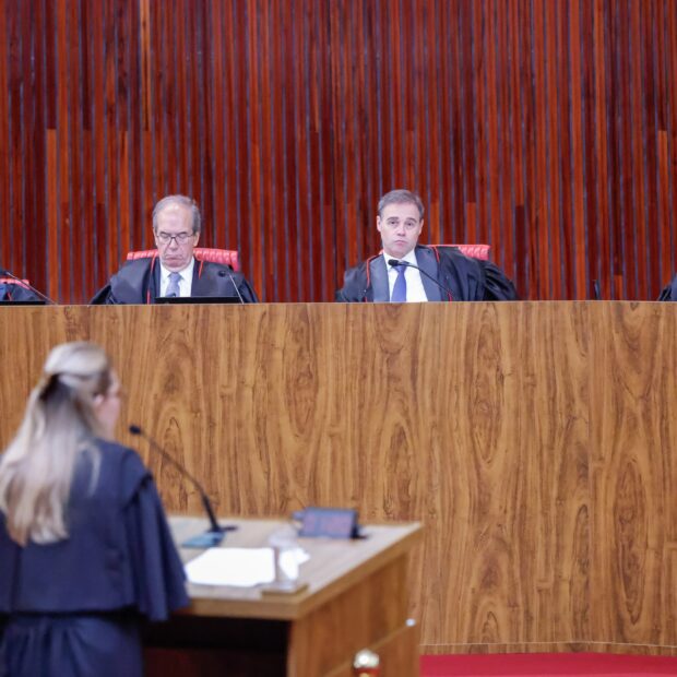 Quatro juízes com togas pretas sentados atrás de uma bancada de madeira enquanto uma pessoa de costas fala ao microfone em um tribunal.