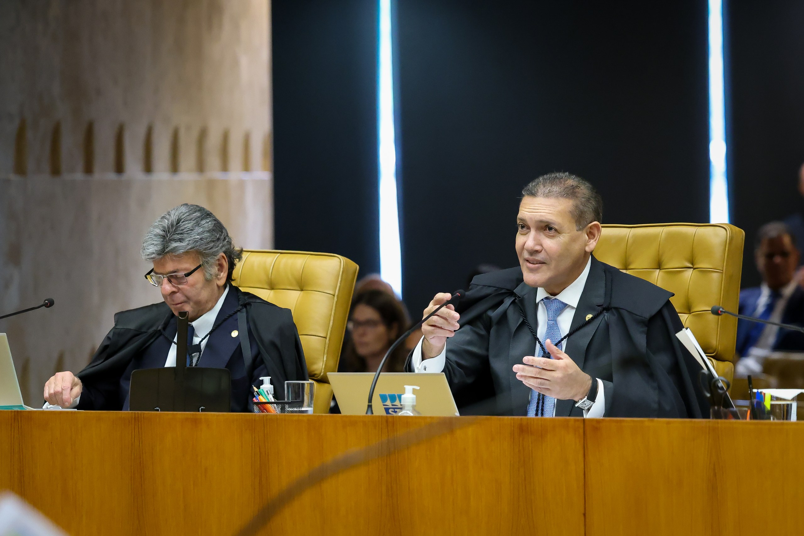 Dois homens com trajes judiciais sentados à mesa em uma sessão de tribunal, com um deles gesticulando enquanto fala. Na foto: Nunes Marques.