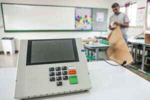 Urna eletrônica de votação sobre uma mesa branca em sala de aula, com um homem ao fundo manuseando papel pardo.