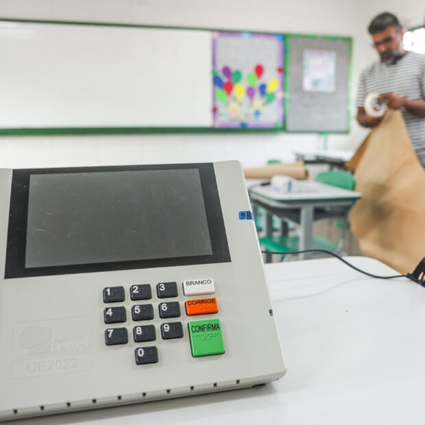 Urna eletrônica de votação sobre uma mesa branca em sala de aula, com um homem ao fundo manuseando papel pardo.